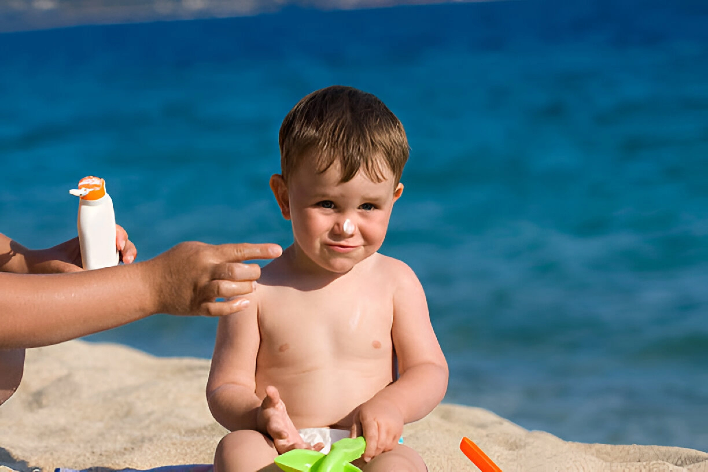 A mother is sitting on the beach in the sun, applying sunscreen to her child's body.
