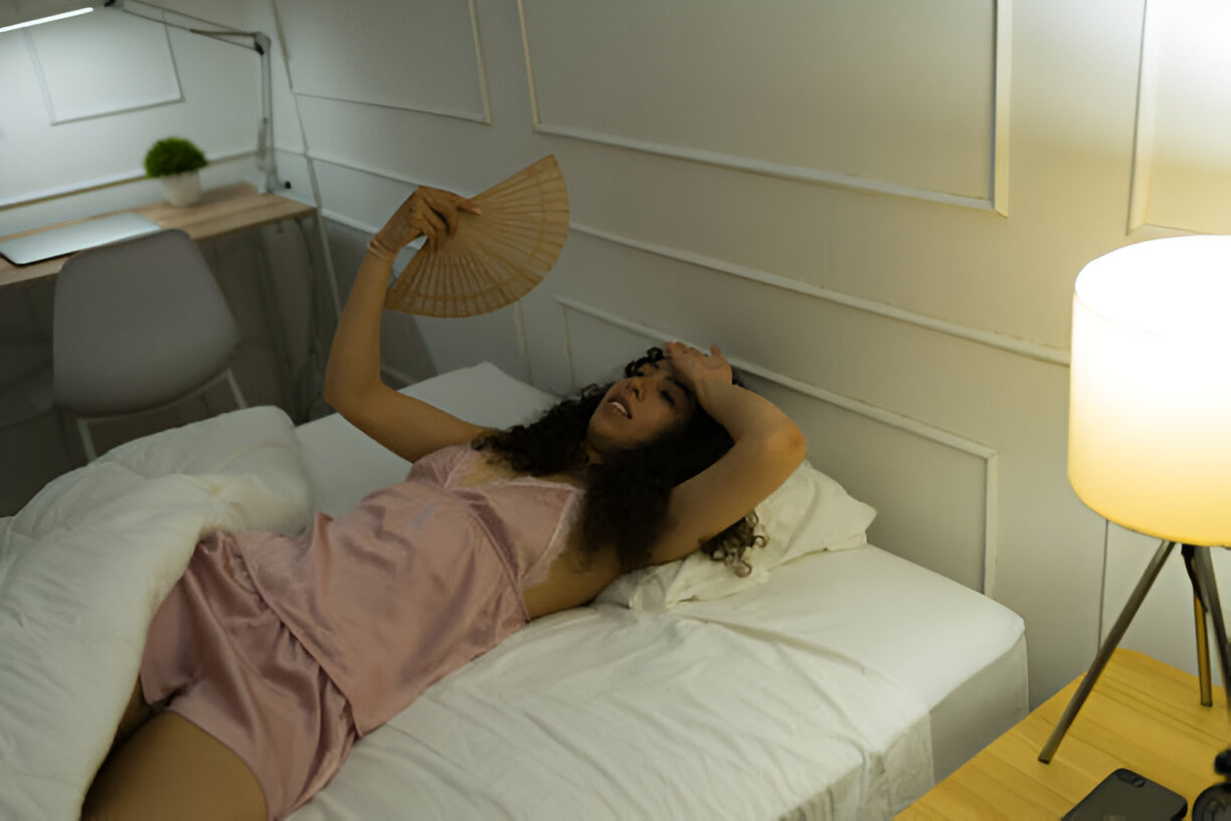 Here, a woman is fanning the air with a hand fan, as she disturbs her sleep due to the heat. how Cooling Mattresses for Hot Sleepers can help prevent discomfort and improve sleep quality.