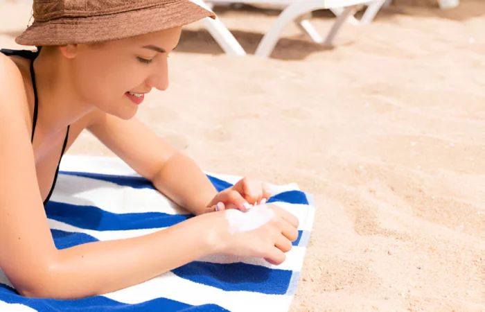 A girl is sitting on the beach in the sun, applying sunscreen to her body.