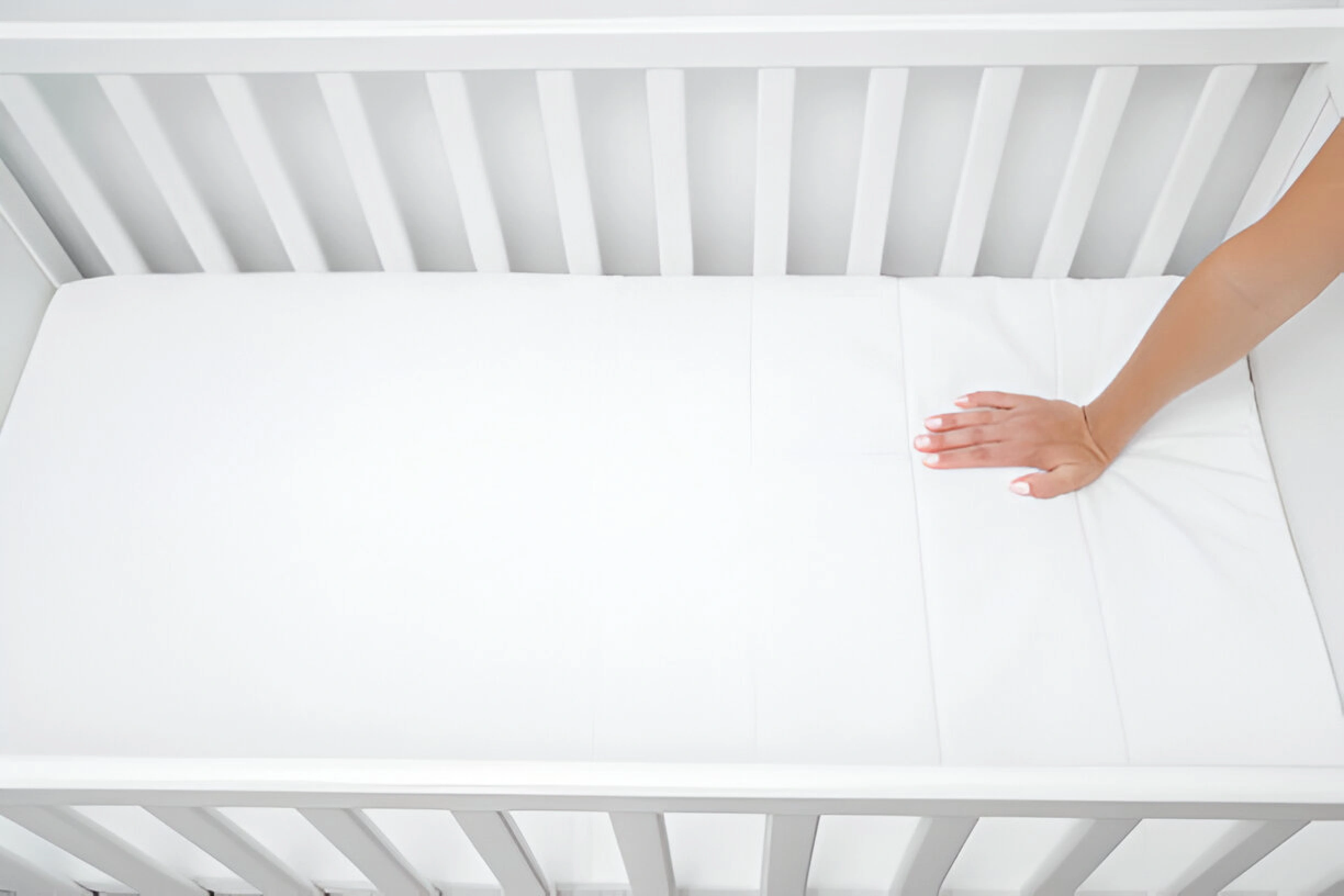 There is a white mattress on a crib, where someone is checking the mattress with their hands.